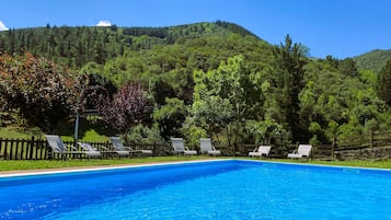 A swimming pool with lounge chairs and a wooden fence, surrounded by lush greenery and hills.
