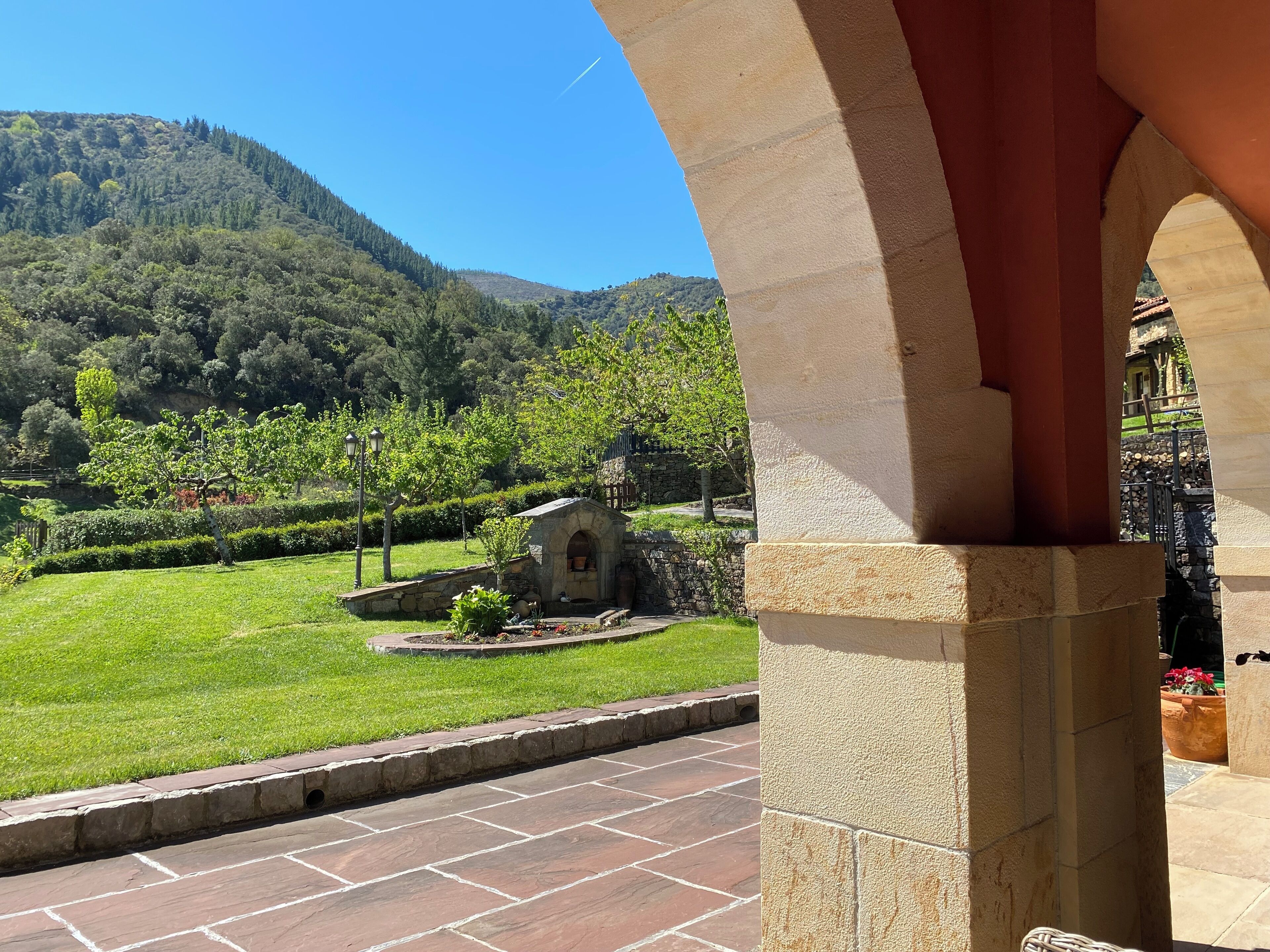 Ein steiner Torbogen führt zu einem Innenhof mit einer kleinen Steinstruktur, umgeben von gepflegtem Rasen und Bäumen, mit einem Berg im Hintergrund.