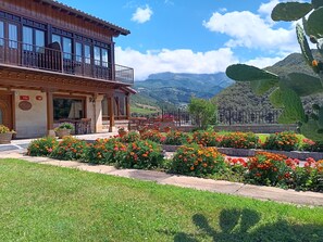 A wooden building with a balcony, surrounded by a garden with marigold flowers, and a mountainous backdrop. - Hotel Finca Malvasia (Cabezon de Liebana)