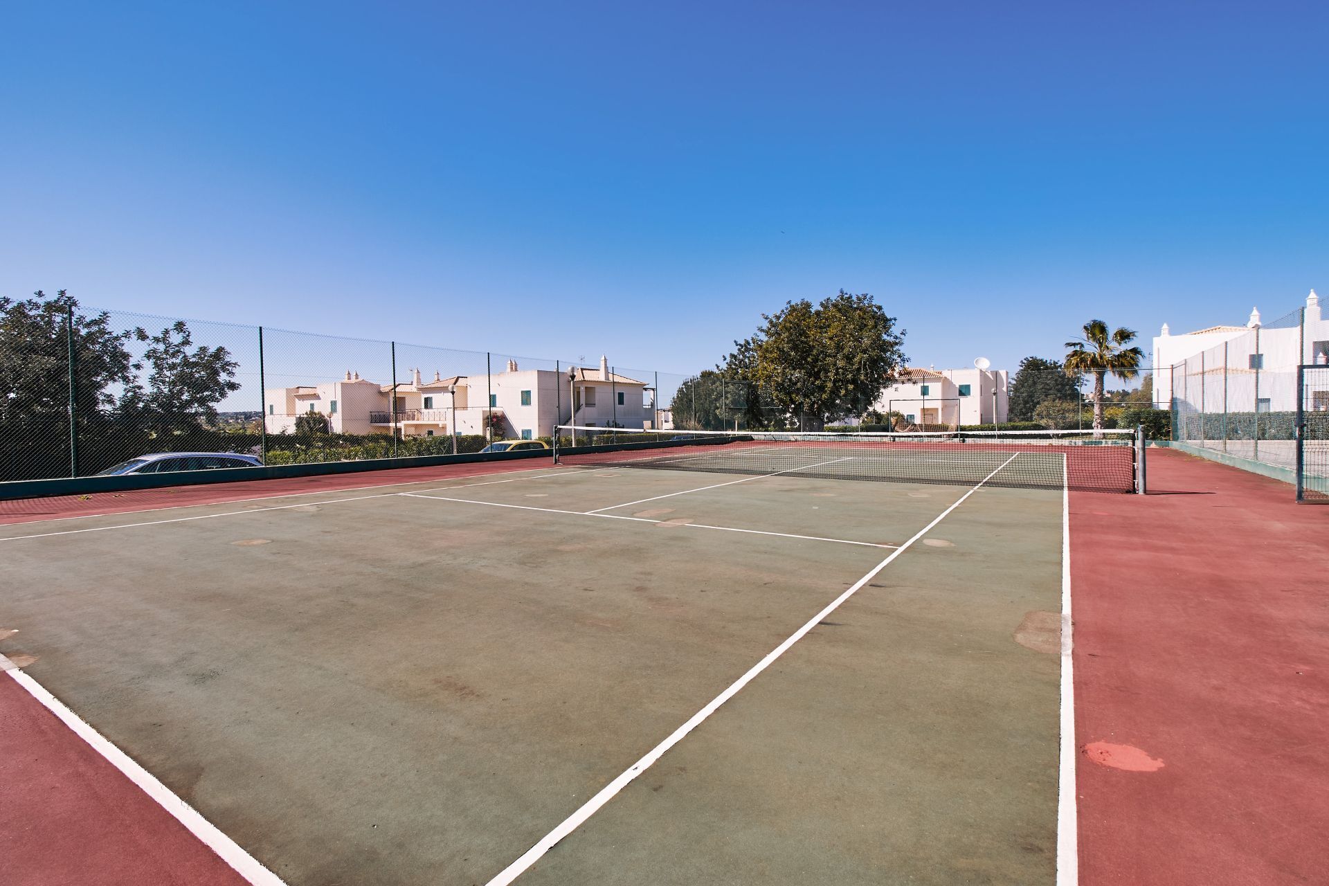 An outdoor tennis court with a net, surrounded by a fence, and buildings in the background.