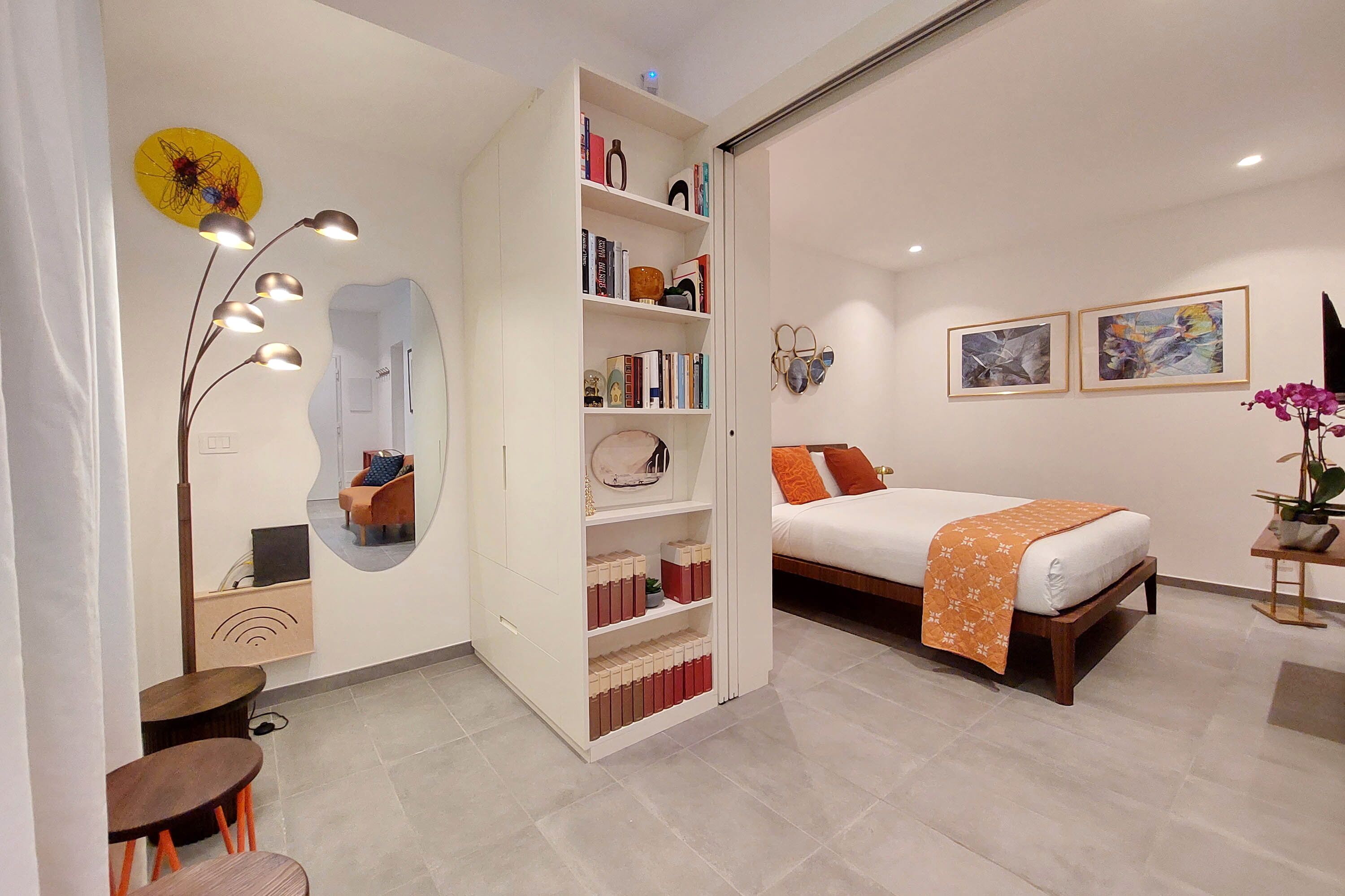 A modern bedroom with a bed, a bookshelf, a mirror, and a small table with a plant.