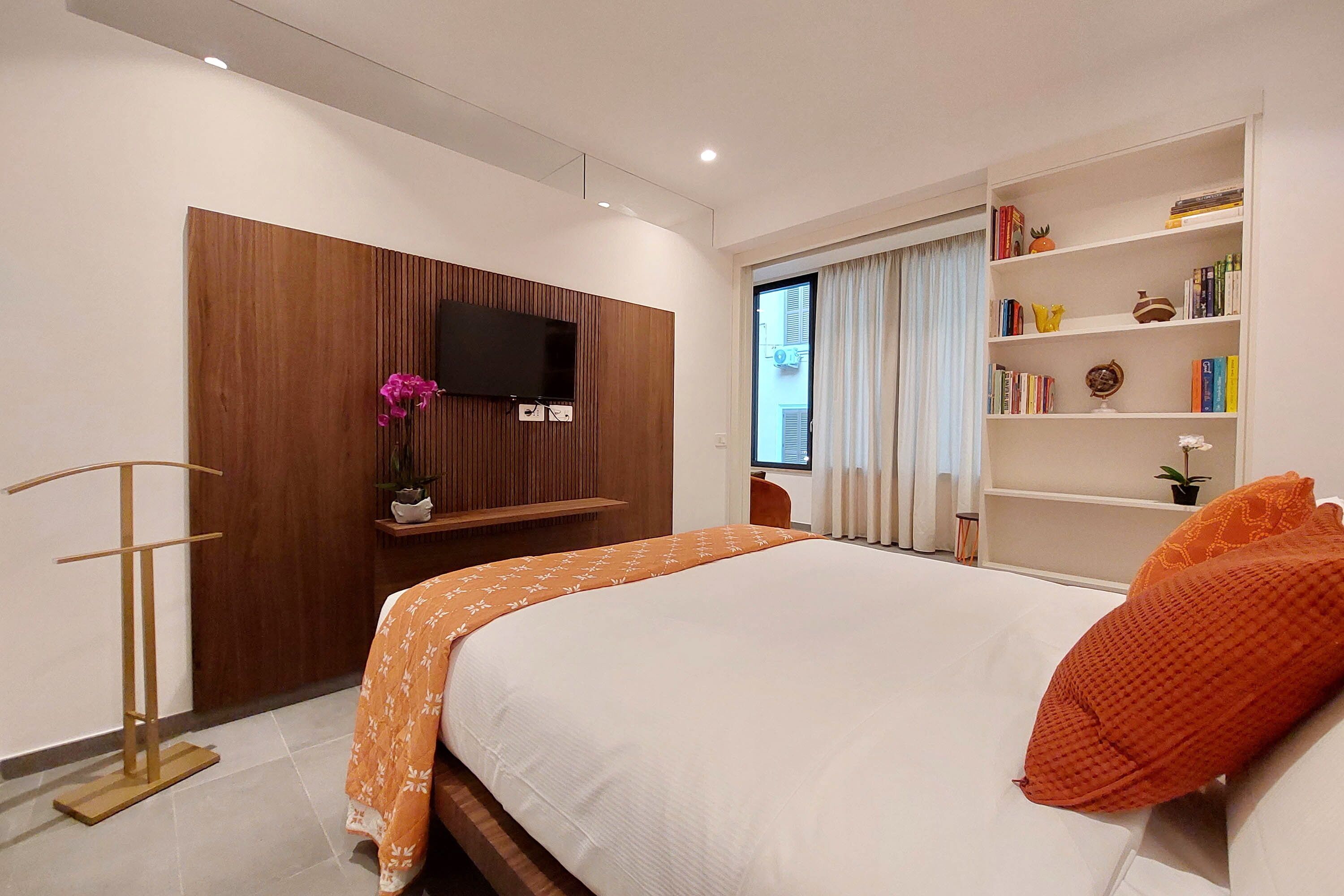 Een moderne slaapkamer met een groot bed, een televisie bevestigd aan een houten paneel, een boekenkast en een raam met gordijnen.