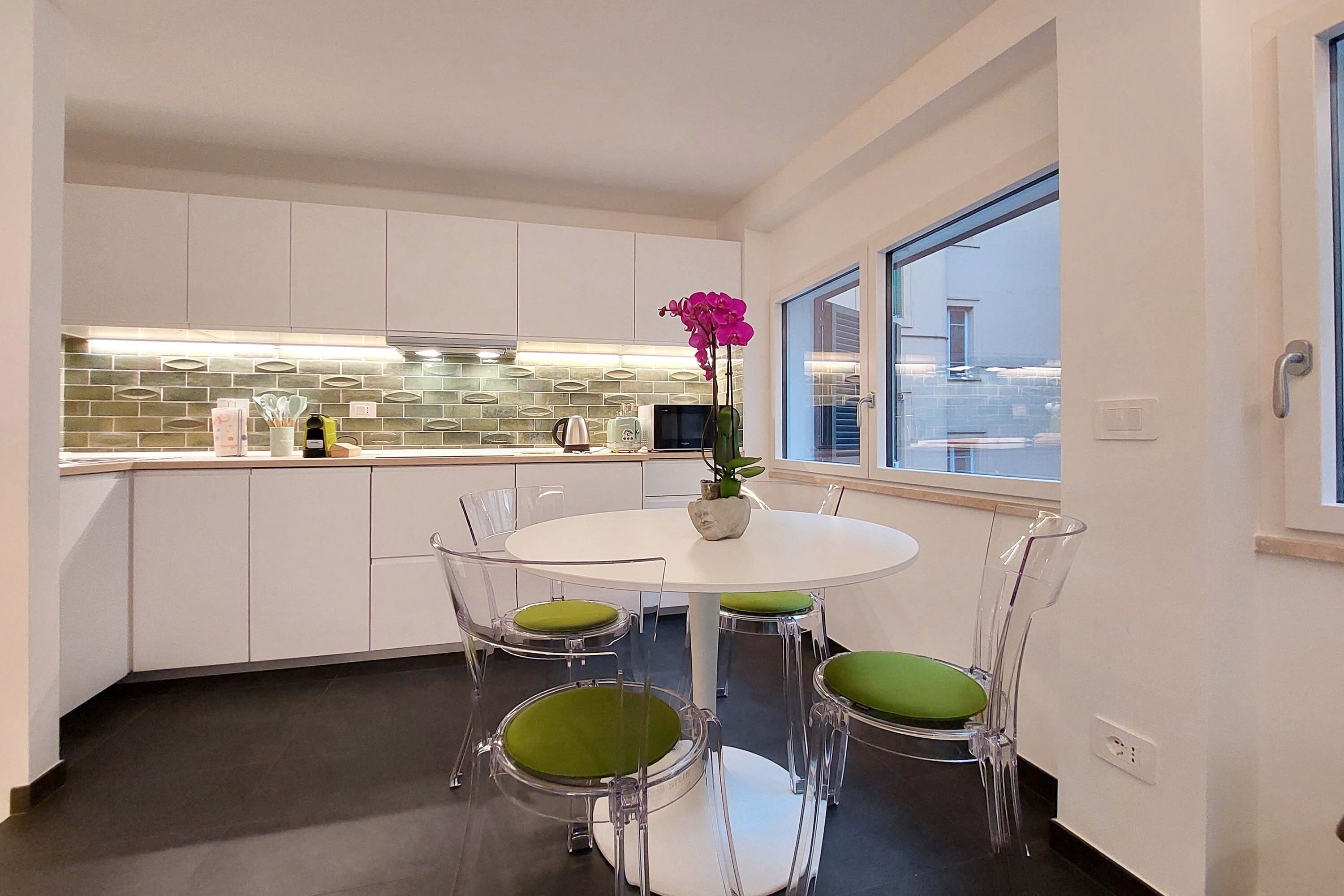 A modern kitchen with white cabinets, a tiled backsplash, and a dining area with a white table and clear chairs with green cushions.