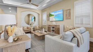 A living room with a sofa, coffee table, and TV. There is a fireplace, a ceiling fan, and a large window with blinds. - 3134 Estero Blvd 5 Bedroom Home by RedAwning (Fort Myers Beach)
