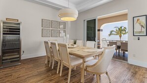 A dining area with a wooden table and chairs, a wine refrigerator, and a large window with a view of palm trees. - 3132 Estero Blvd 5 Bedroom Home by RedAwning (Fort Myers Beach)