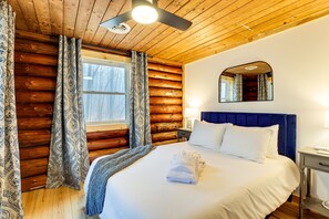 A log cabin room with a bed, a nightstand, a mirror, and a window with curtains. - Secluded Naples Cabin w/ Deck & Stream Views (Naples)