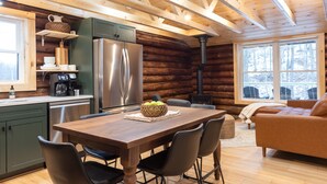 L’intérieur d’une cabane en rondins comprend une table à manger en bois, des chaises, un canapé, une cuisine avec un réfrigérateur et une grande fenêtre.