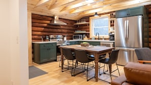 A modern kitchen with wooden beams, a stainless steel refrigerator, and a dining area with a table and chairs. - Secluded Naples Cabin w/ Deck & Stream Views (Naples)