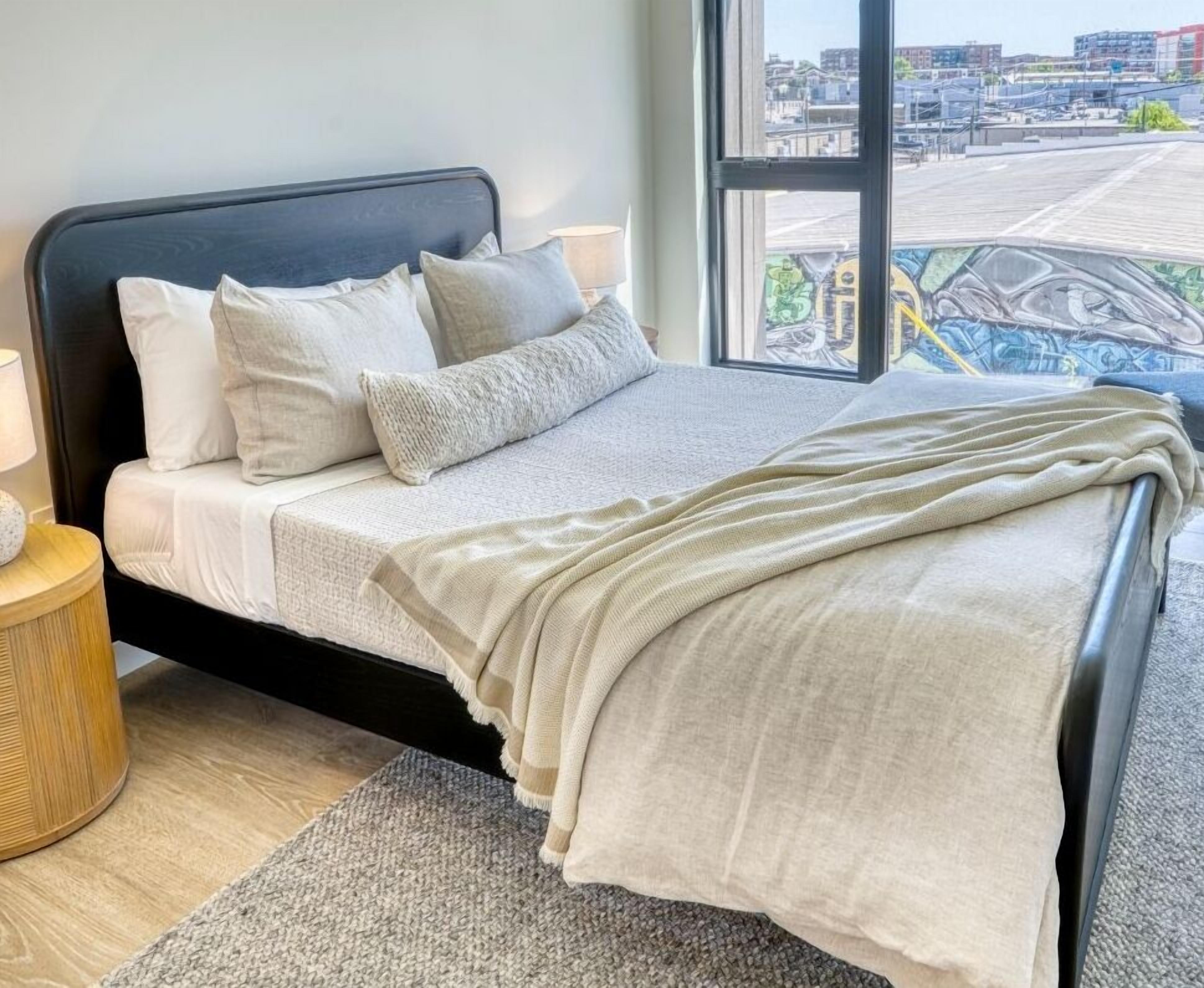 A modern bedroom with a large bed, a nightstand, a lamp, and a view of the city through a large window.