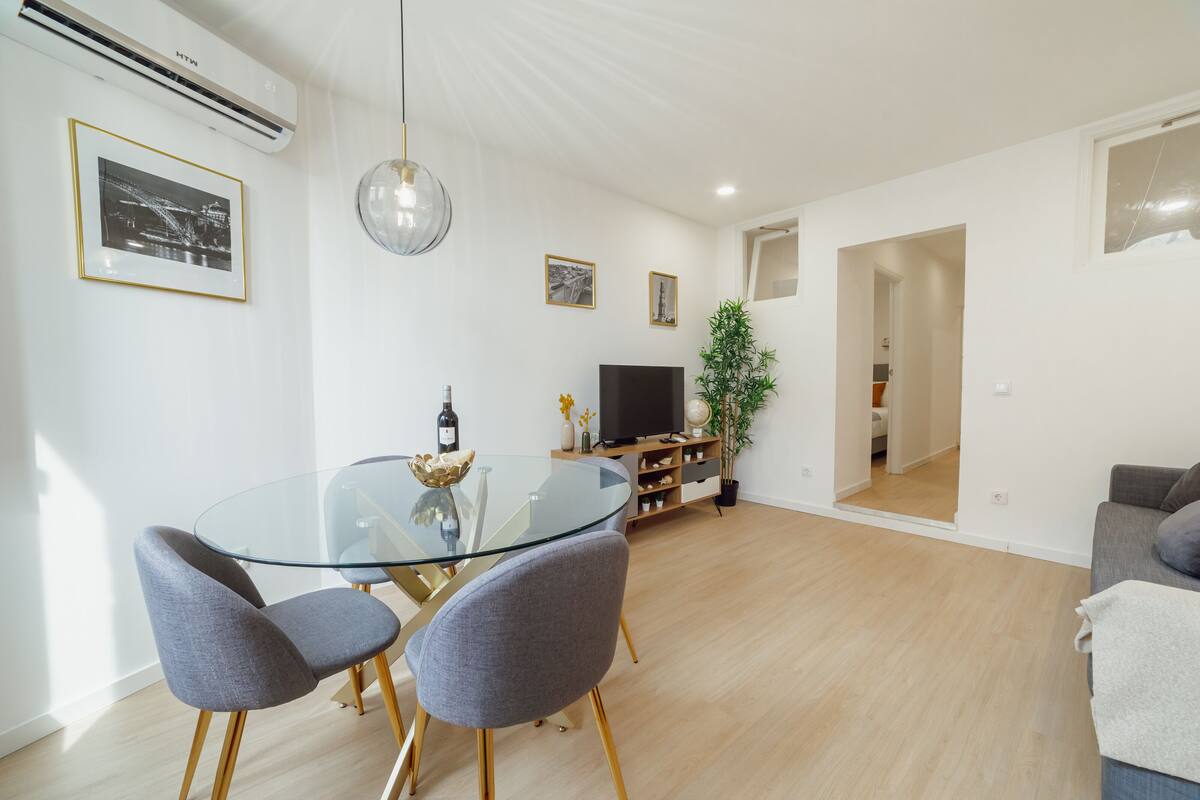 Un salon moderne avec une table à manger en verre, des chaises recouvertes d’un tissu gris, un téléviseur à écran plat et une plante en pot.