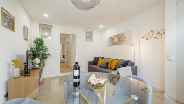 Ein modernes Wohnzimmer mit einem Glastisch, grauen Sofas und einer großen Topfpflanze.