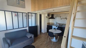 Een ruimtelijk ingerichte leefruimte met een bank, een klein rond tafeltje, een kitchenette en een houten lattenplafond.