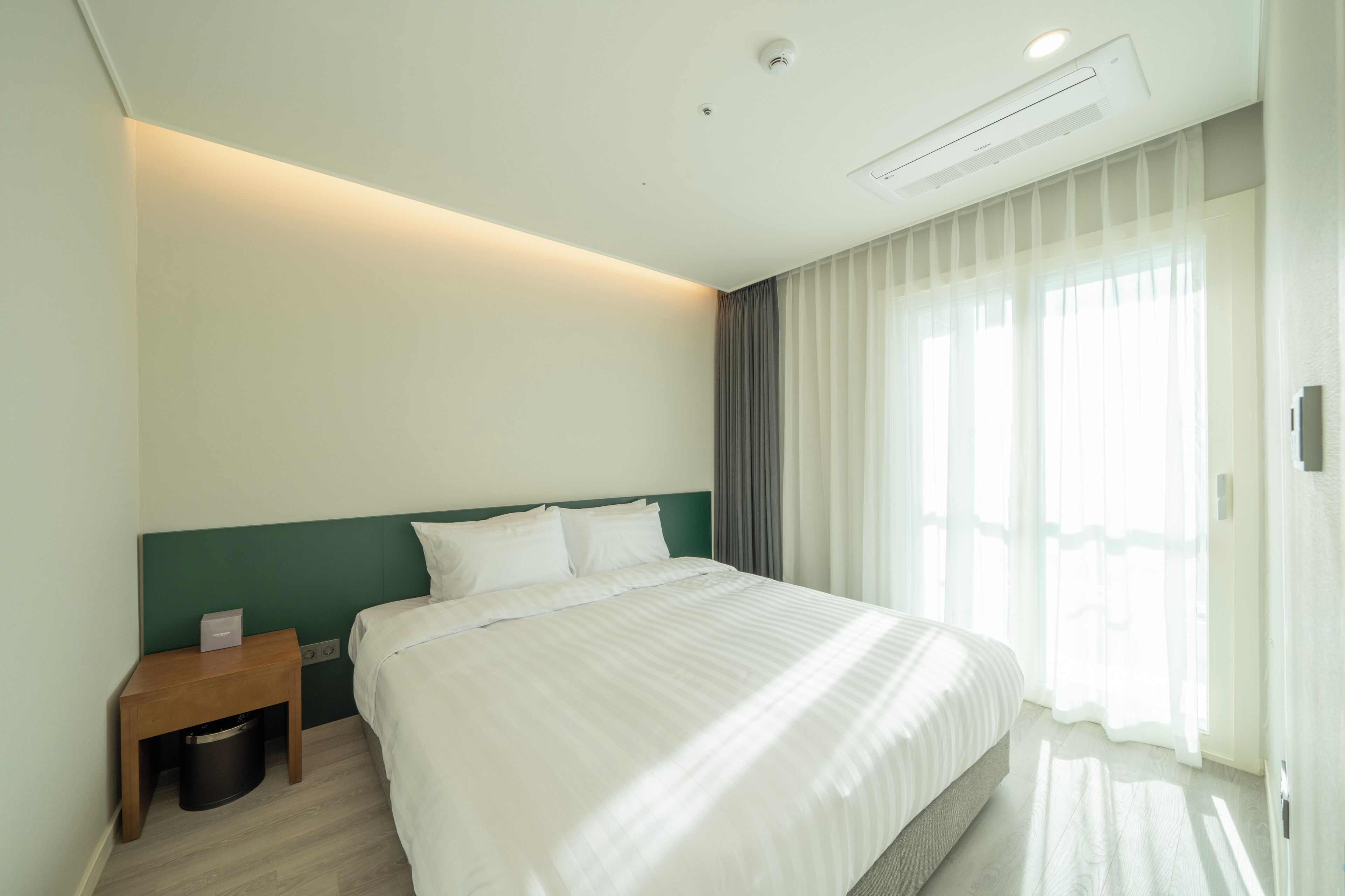 A hotel room with a bed, a nightstand, a trash bin, a window with curtains, and an air conditioning unit.