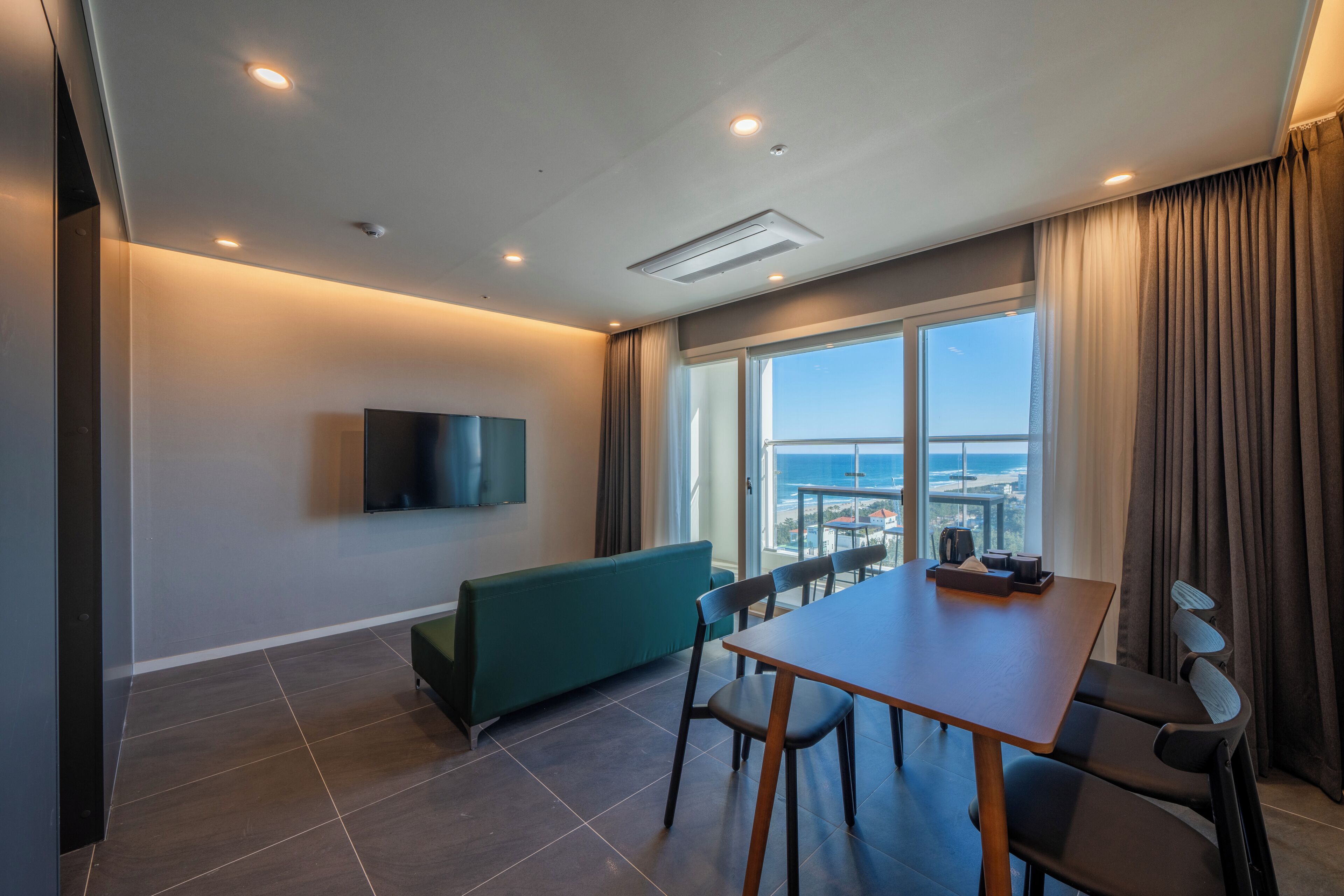 Une chambre d’hôtel moderne dotée d’un coin repas, d’un téléviseur à écran plat fixé au mur, d’une grande fenêtre donnant sur l’océan et d’un plafond équipé d’un éclairage encastré.