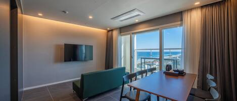 Une chambre d’hôtel moderne dotée d’un coin repas, d’un téléviseur à écran plat fixé au mur, d’une grande fenêtre donnant sur l’océan et d’un plafond équipé d’un éclairage encastré.