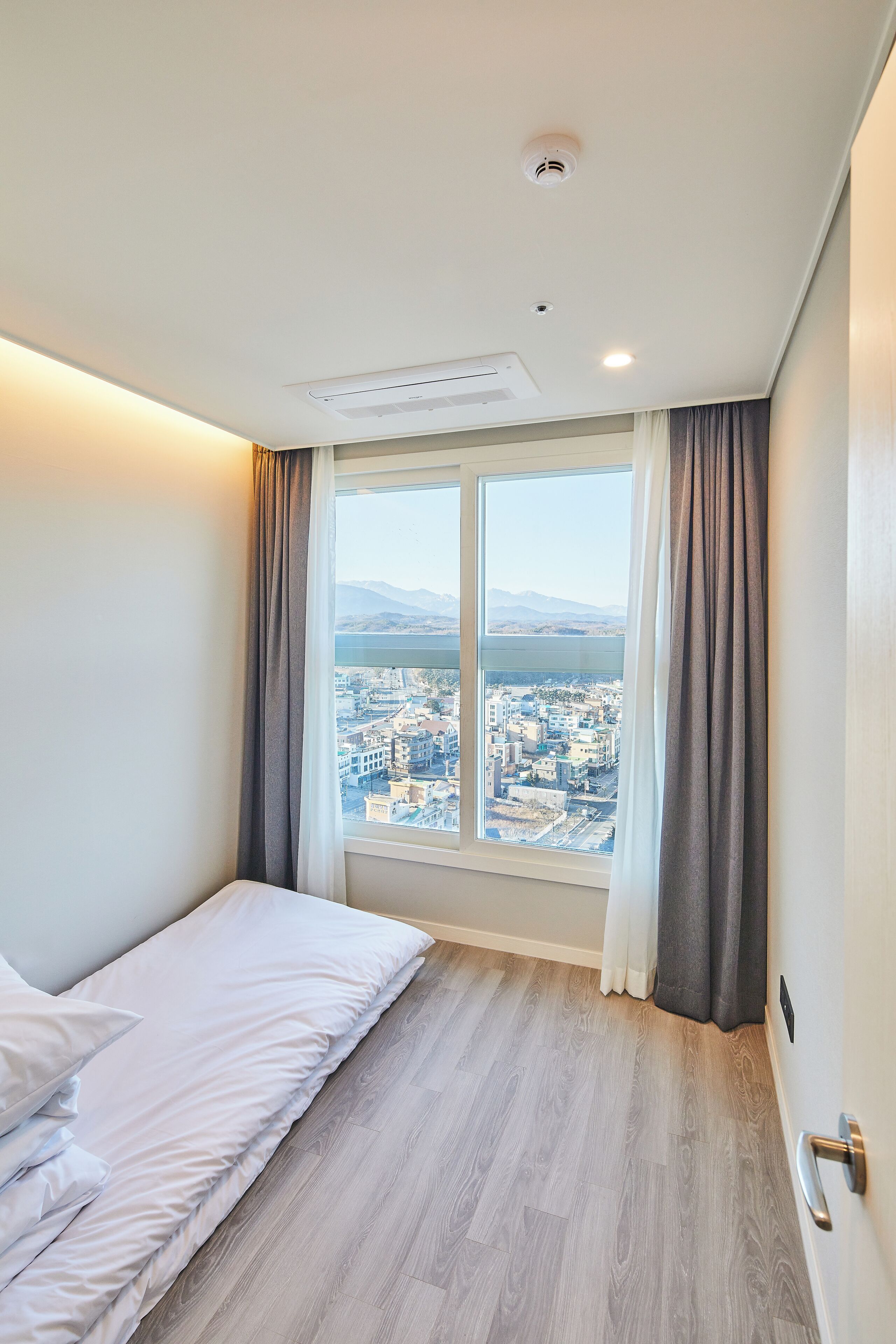 A bedroom with a bed, a window with curtains, a view of a cityscape, and a ceiling with a smoke detector.