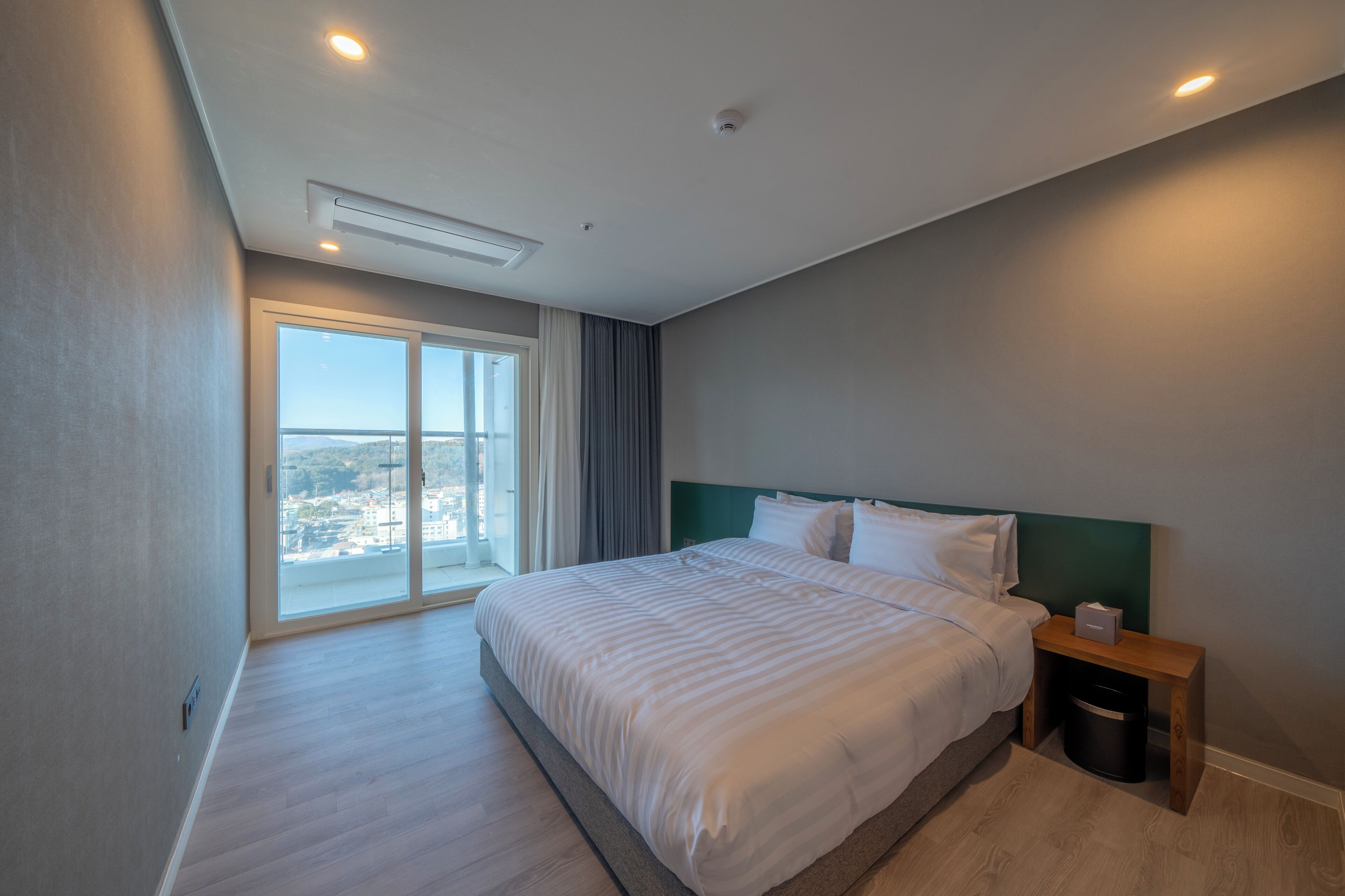 A modern hotel room with a large bed, a nightstand, a trash bin, and a view of the city through a large window.