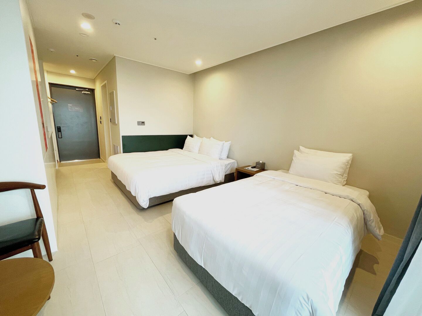 Ein Hotelzimmer mit zwei Betten, einem Holzstuhl und einer Tür zu einem Flur.
