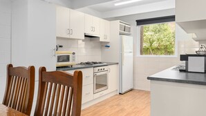 Eine moderne Küche mit weißen Schränken, einer Mikrowelle, einem Herd und einem Kühlschrank. Ein Esstisch mit Stühlen und ein Fenster mit Blick auf Bäume.