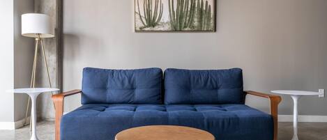 A modern living room with a blue sofa, two white side tables, a round wooden ottoman, and a cactus painting on the wall.