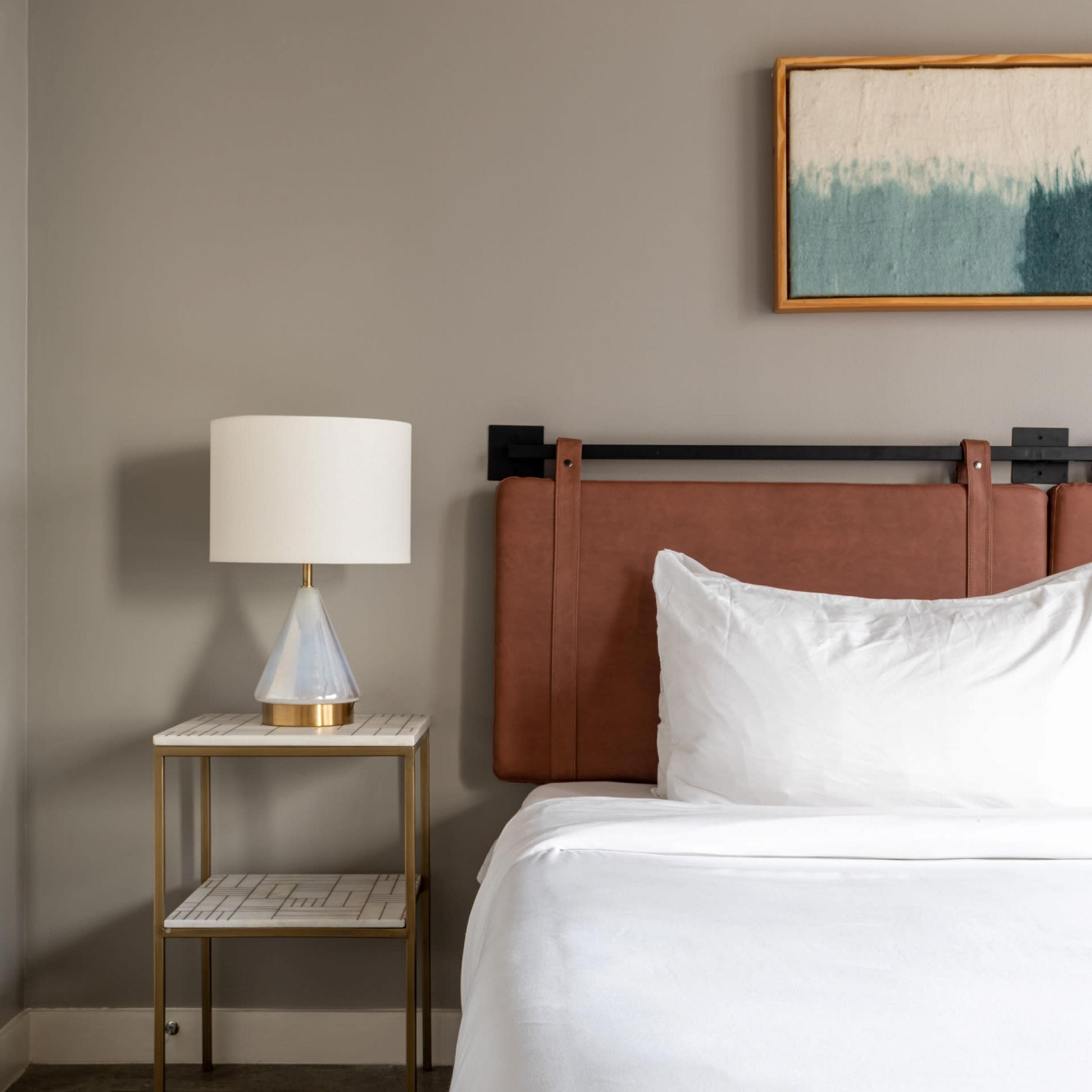 A hotel room with a bed, a bedside table with a lamp, and a painting on the wall.