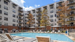 An outdoor swimming pool with lounge chairs and umbrellas, surrounded by multi-story residential buildings with balconies.