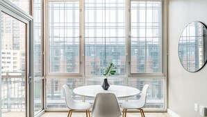 A modern dining area with a round table and four chairs, large windows with a city view, and a round mirror on the wall.