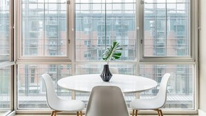 A modern dining area with a round table and four white chairs, a vase with a plant, and large windows offering a view of a cityscape. - Austin Gorgeous 1BD 1BA Downtown Apartment (Austin)