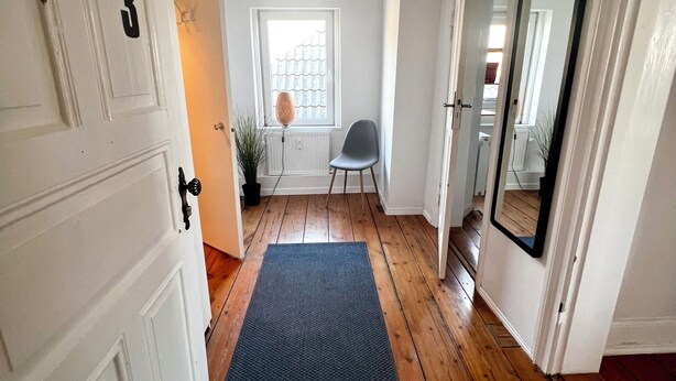A hallway with wooden floors, a blue mat, a radiator, a chair, and a mirror. - Ferienwohnung Apartment No. 9 in Husum für bis zu 2 Personen (Husum)