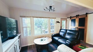 Un espace de vie compact comprenant un téléviseur, un canapé en cuir noir, un fauteuil blanc et une petite table ronde. De grandes fenêtres offrent une vue sur l’extérieur.