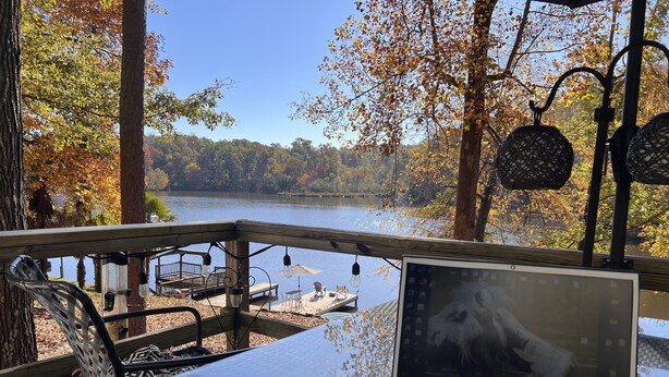 Un ordinateur portable posé sur une table en verre, avec vue sur un lac et des arbres aux couleurs automnales.