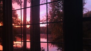 Une vue à travers une fenêtre à carreaux, montrant un coucher de soleil sur une étendue d’eau bordée d’arbres et d’un quai.