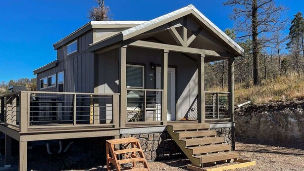 A modern cabin with a covered deck, a staircase, and a view of the surrounding forest. - Bear Hug Cabin (Ruidoso Downs)