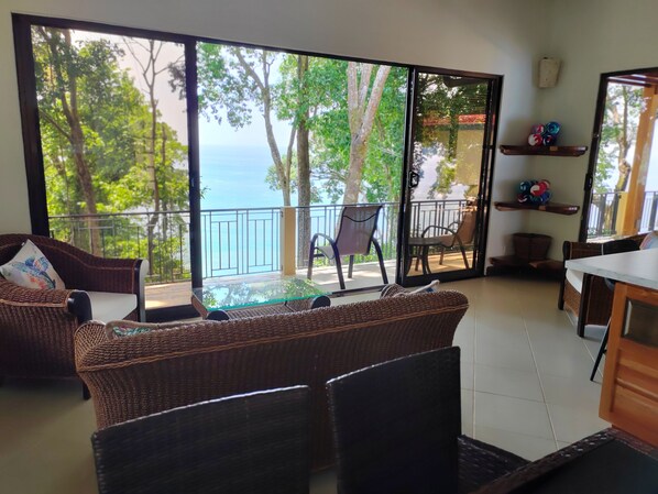 A room with a large sliding glass door leading to a balcony with a view of the sea and trees. There are wicker furniture pieces, a small table, and a shelf with decorative items. - Ocean view Casa Gauguin.
Forget your worries in this spacious and serene space. (Playa dominicalito)