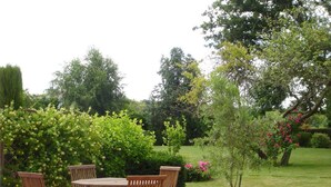 Un patio exterior con una mesa redonda y cuatro sillas de madera, rodeado de abundante follaje y plantas en flor.