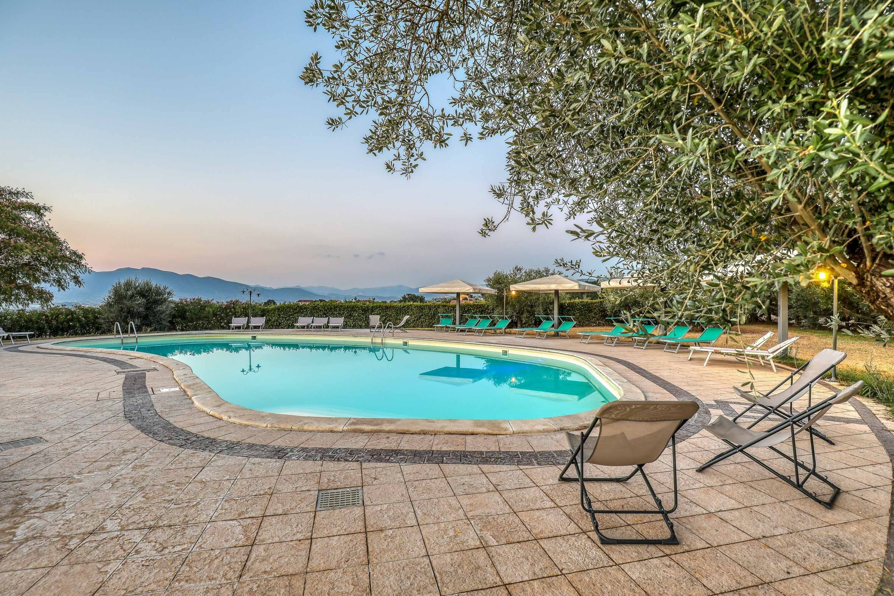 A swimming pool with lounge chairs and umbrellas, surrounded by greenery and mountains in the background.