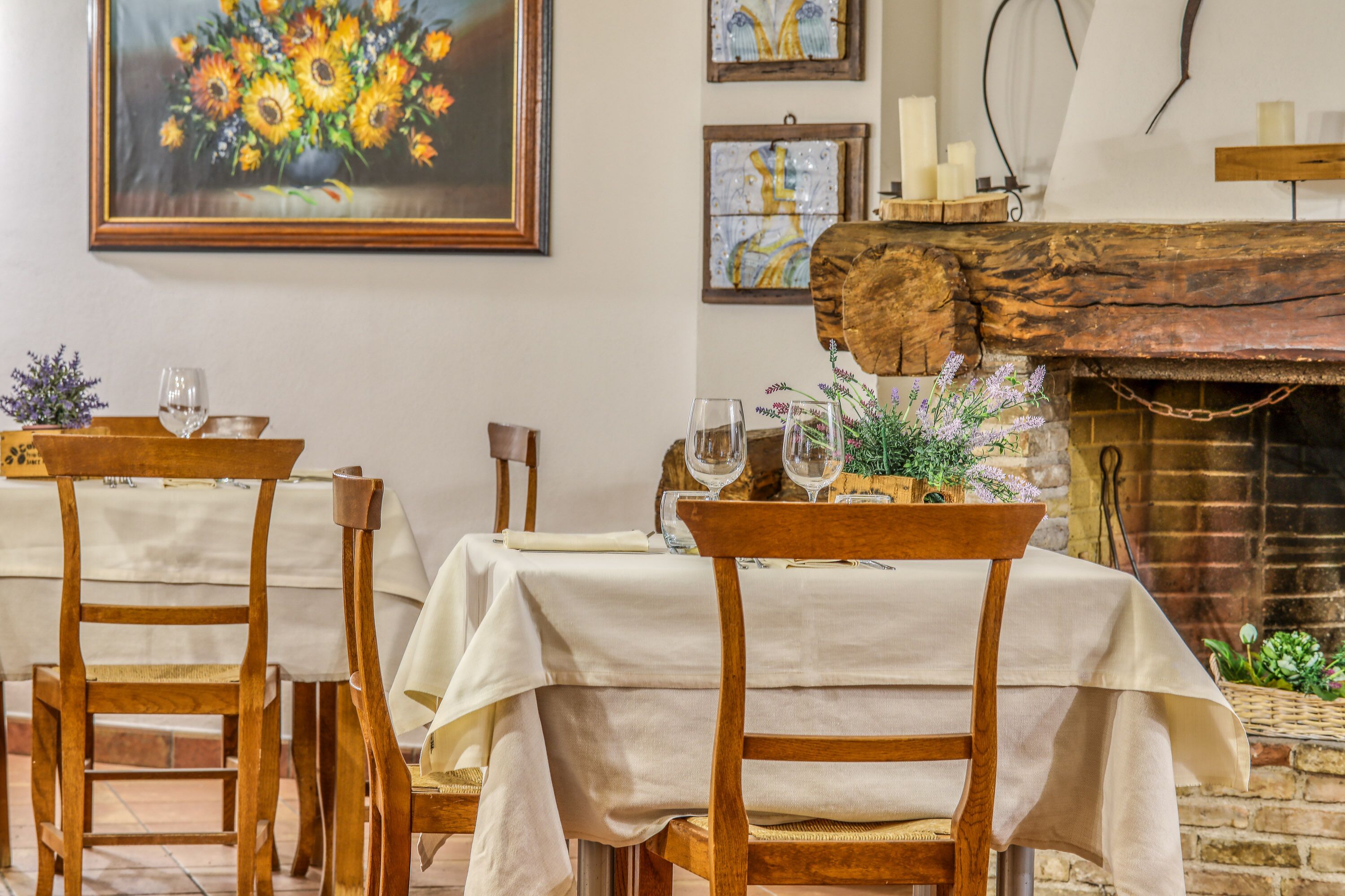 Un comedor con sillas y mesas de madera cubiertas con manteles blancos, una chimenea rústica y cuadros en las paredes.
