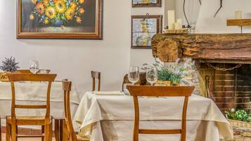 Un comedor con sillas y mesas de madera cubiertas con manteles blancos, una chimenea rústica y cuadros en las paredes.