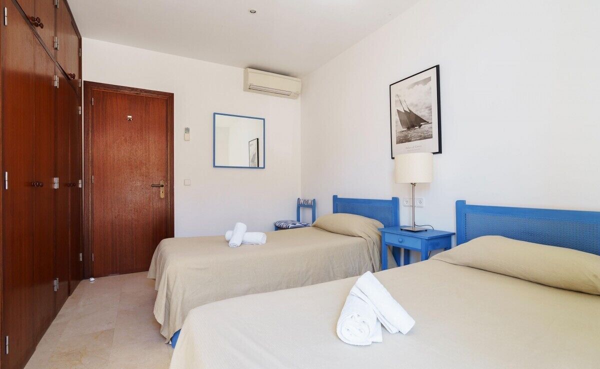 A hotel room with two beds, a wardrobe, a mirror, a framed picture, and a bedside table with a lamp.