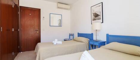 A hotel room with two beds, a wardrobe, a mirror, a framed picture, and a bedside table with a lamp.