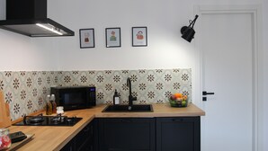Une cuisine compacte équipée d’un four à micro-ondes, d’un évier et de meubles de couleur noire. Trois tableaux encadrés ornent le mur, un panier de fruits est posé sur le comptoir et un luminaire noir suspendu éclaire l’ensemble.