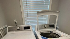 Une buanderie équipée d’un lave-linge et d’un sèche-linge à chargement frontal, d’une prise de courant et d’une fenêtre avec des stores.