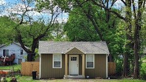 Une petite maison de plain-pied, avec une porte d’entrée et deux fenêtres, entourée d’arbres et d’une cour gazonnée.