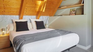 A bedroom with a sloped ceiling, a bed with white and black bedding, a nightstand, a lamp, and a shelf with books. - Les Marmottes by FeelFree Rentals (Vielha)