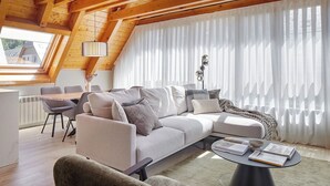 A modern loft-style living room with a high ceiling, a large window with sheer curtains, a grey sofa set, a round coffee table, and a dining area with a table and chairs. - Les Marmottes by FeelFree Rentals (Vielha)