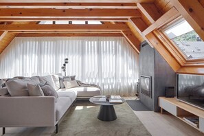 Un dormitorio tipo loft moderno con techo inclinado, un ventanal con cortinas translúcidas, televisor integrado y una zona de estar acogedora con sofá y mesa de centro.