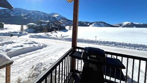 A snow-covered mountain landscape with a clear sky, a wooden balcony with a grill, and a view of houses in the distance. - Elk Valley- Views for days (Crested Butte)