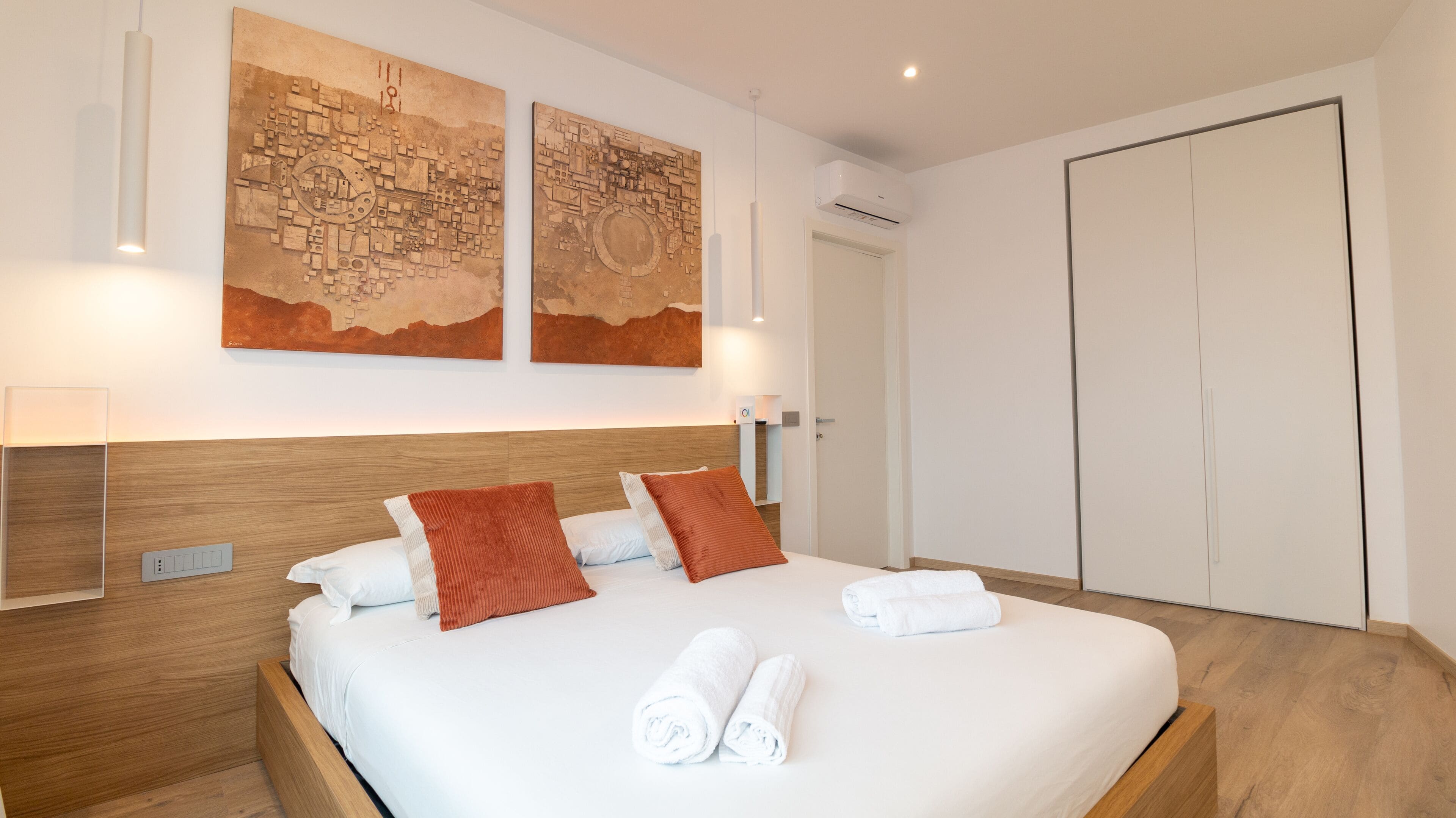 A modern hotel room with a wooden headboard, a large bed with white linens, two red pillows, and two rolled towels. There are two abstract paintings on the wall above the bed, and a white wardrobe to the right.