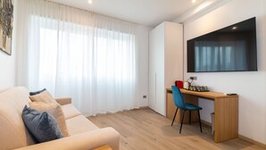 A modern living room with a beige sofa, a wooden desk, a blue chair, and a large flat-screen TV mounted on the wall. - Galleria Roma Art Guesthouse (Oristano)
