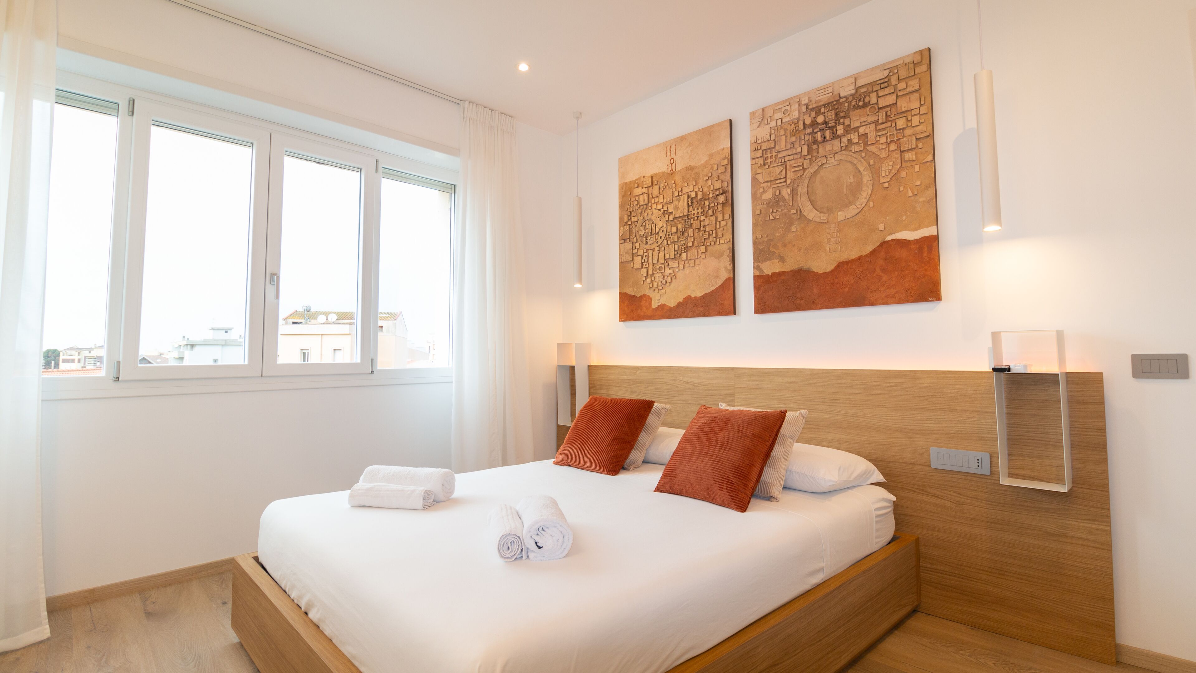 Un lit bien fait, avec des oreillers blancs et rouges, une tête de lit en bois et deux lampes fixées au mur.
